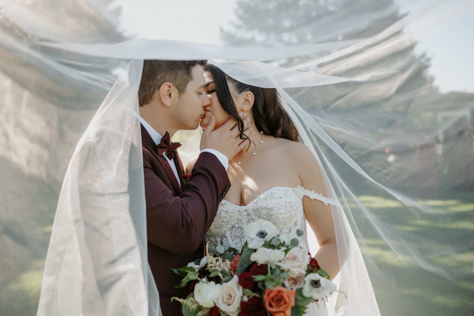 The groom is holding his bride by the chin and they are sharing a kiss underneath her veil.