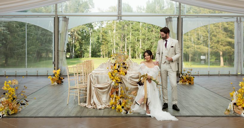Elegant bride and groom next to a stunning rectangular table draped with champagne satin and overloaded with bold yellow arrangements.