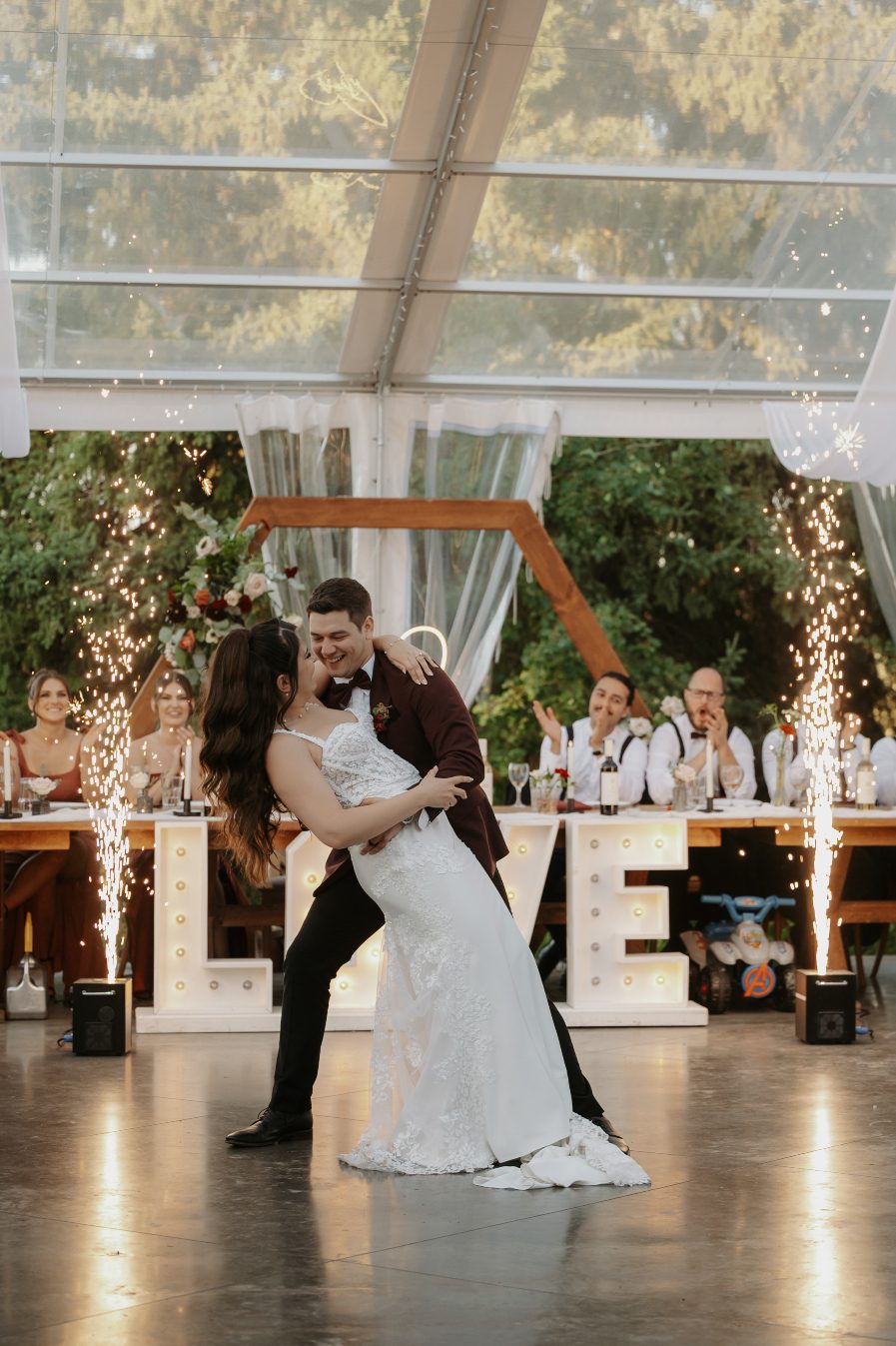The bride and groom sharing a dramatic dip during their first dance as indoor sparklers ignite on each side, with the head table and "LOVE" sign behind them.