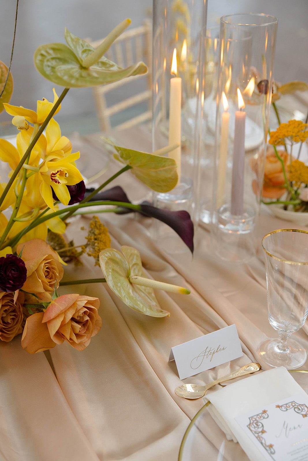 Close up of contemporary yellow wedding florals, including orchids, anthurium and caramel coloured roses arranged alongside tall taper candles on champagne satin linens.