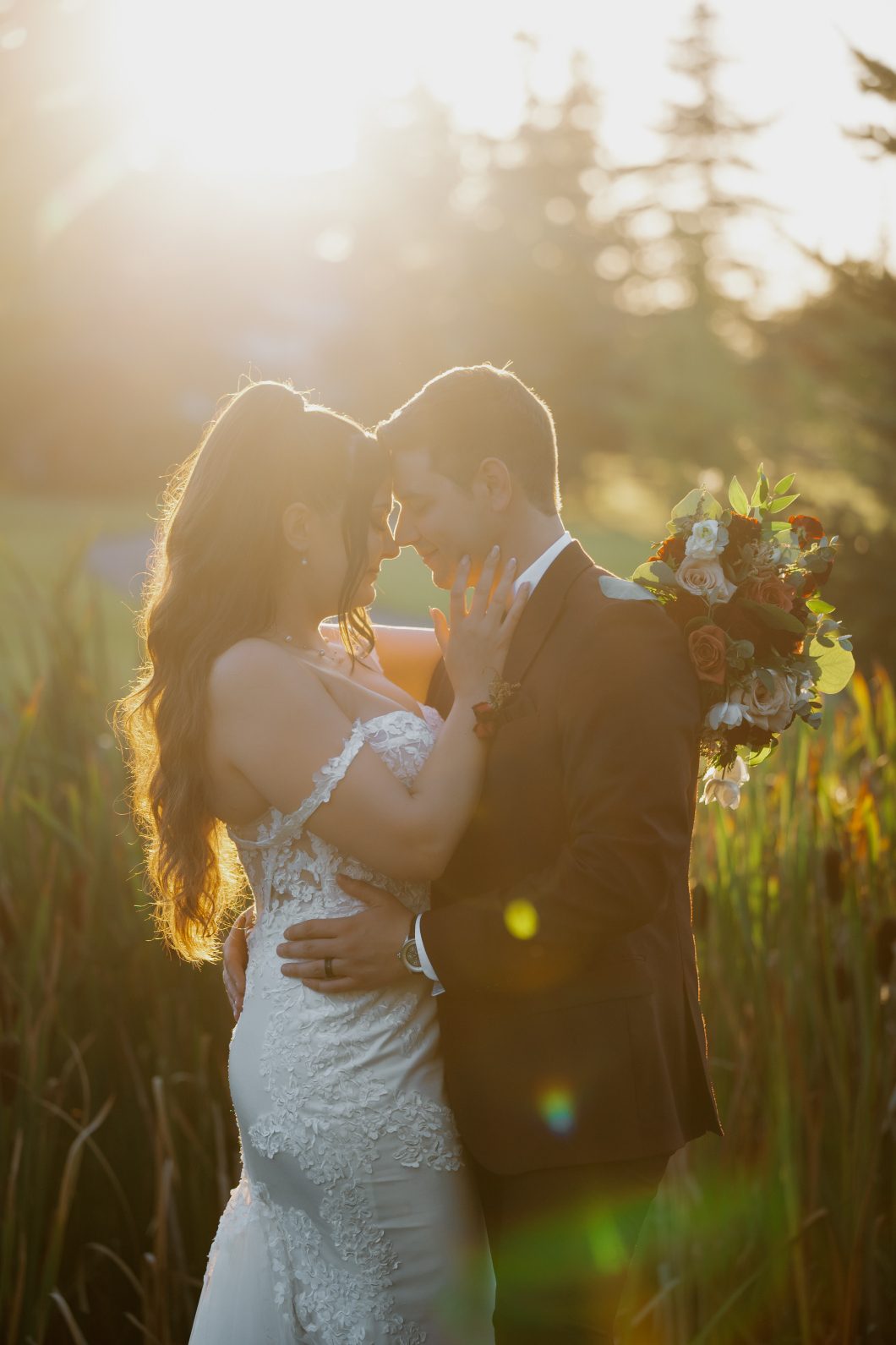 Bride and groom embracing in warm sunset light, with the bride's lace gown glowing and her bouquet held behind the groom's shoulder.