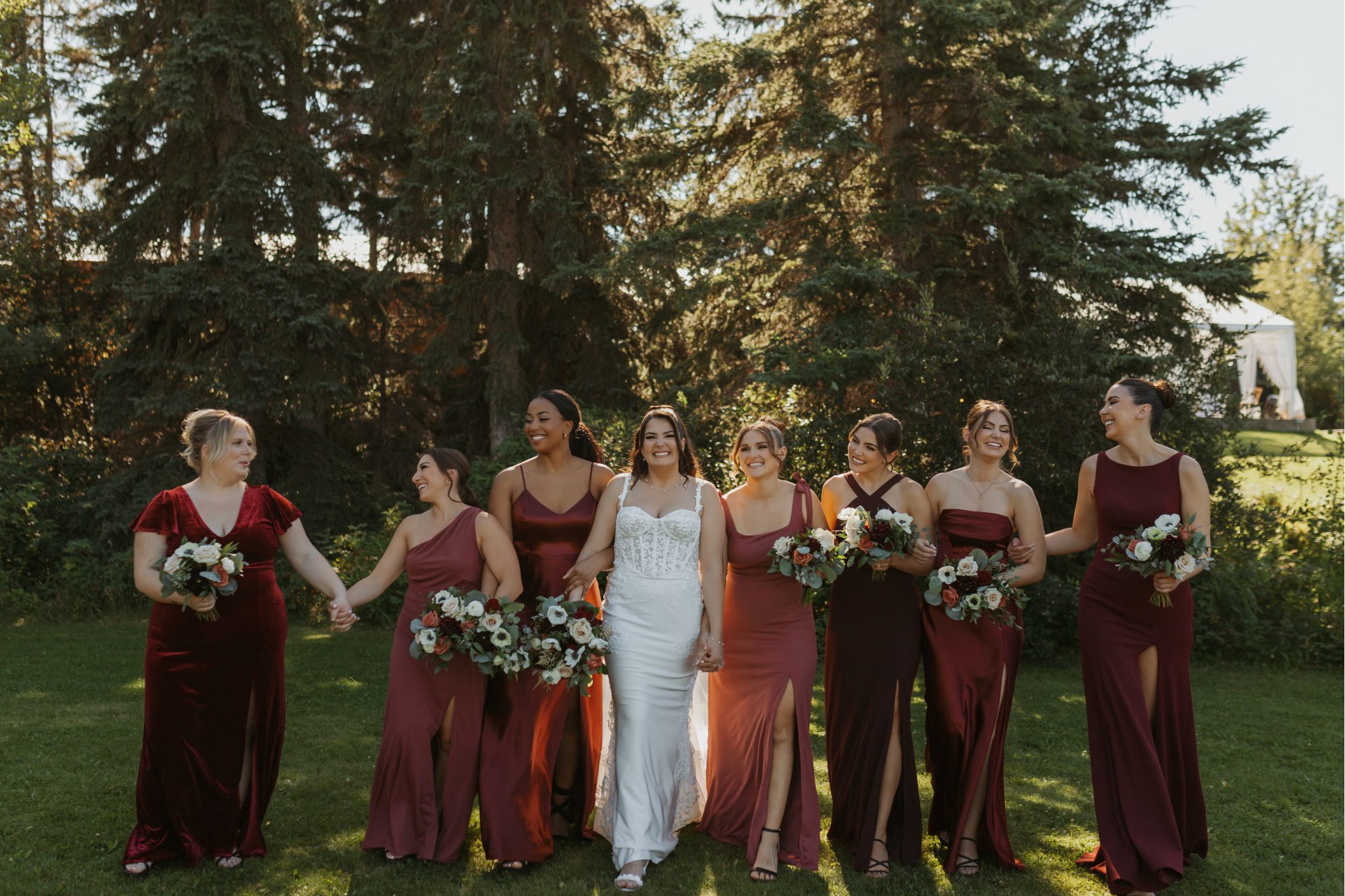 Bride and bridesmaids walking together on a sunny lawn wearing assorted burgundy seasonal floral bouquets.
