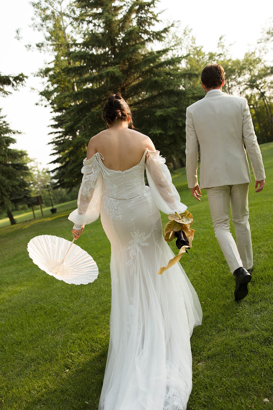 Bride and groom walking through a sunlit grassy field, the bride holding a white parasol and a modern sculptural bouquet.