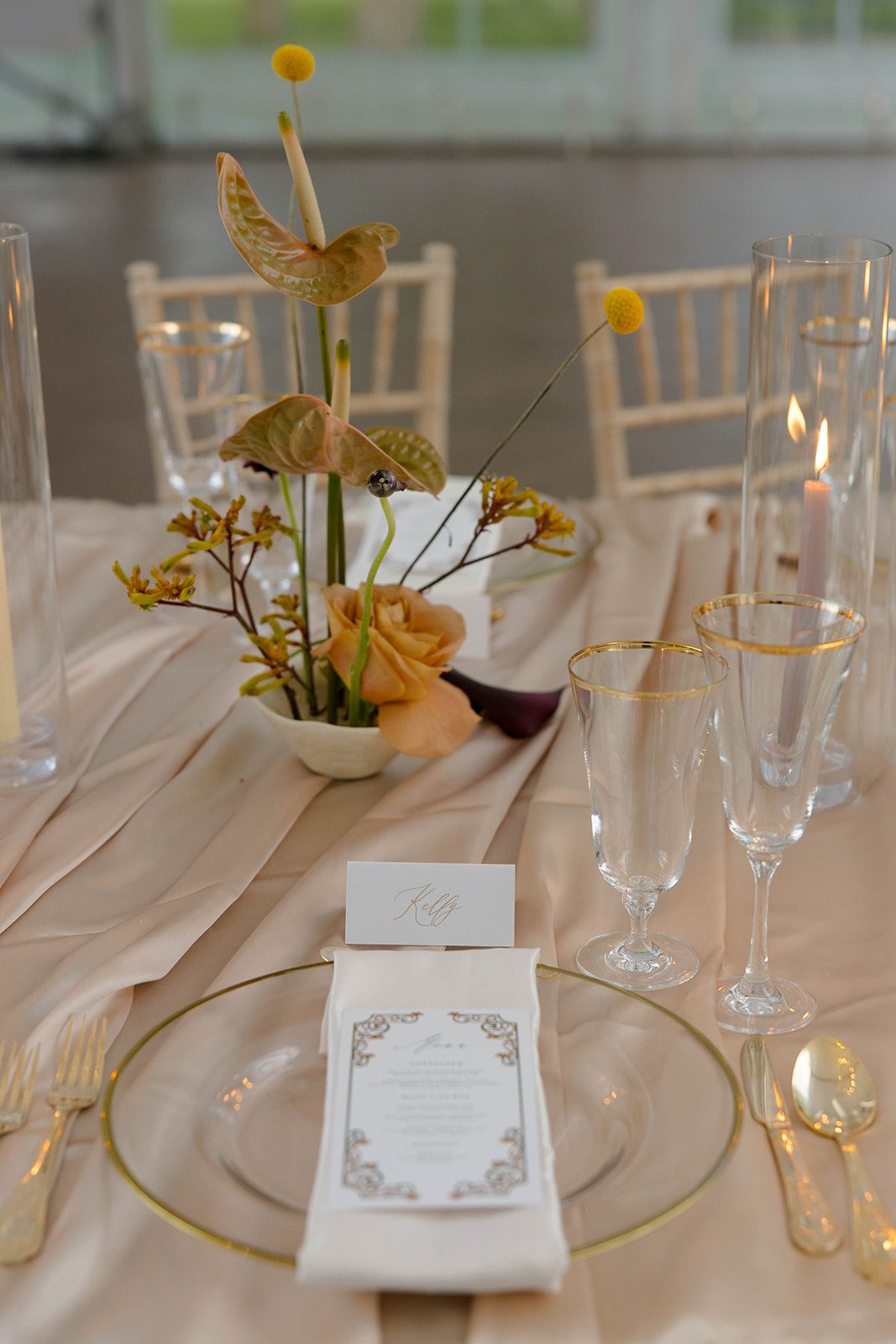 Minimalist ikebana inspired bowl arrangement of anthurium, caramel roses, deep plum calla lilies, and billy buttons besides a gold rimmed place setting and personalized name card.