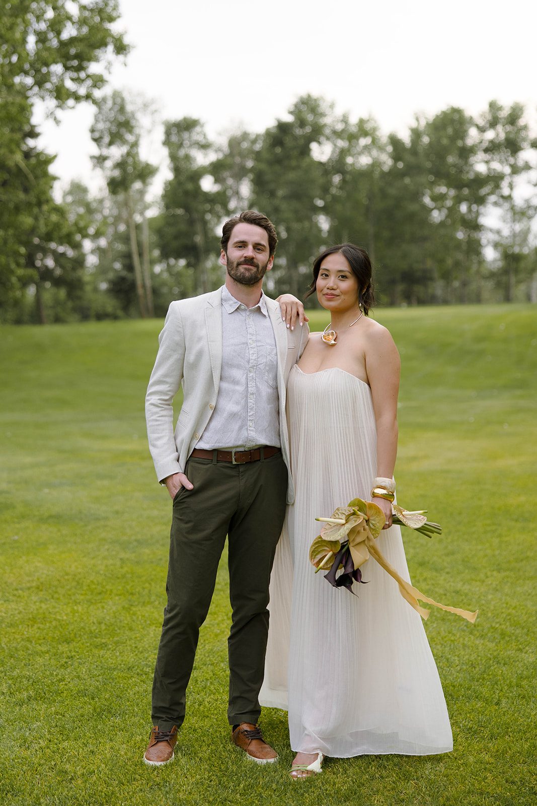 Couple standing together on a lush green lawn, the bride in a pleated cream gown with sculptural jewelry and bouquet, the groom in a light blazer and dark trousers.