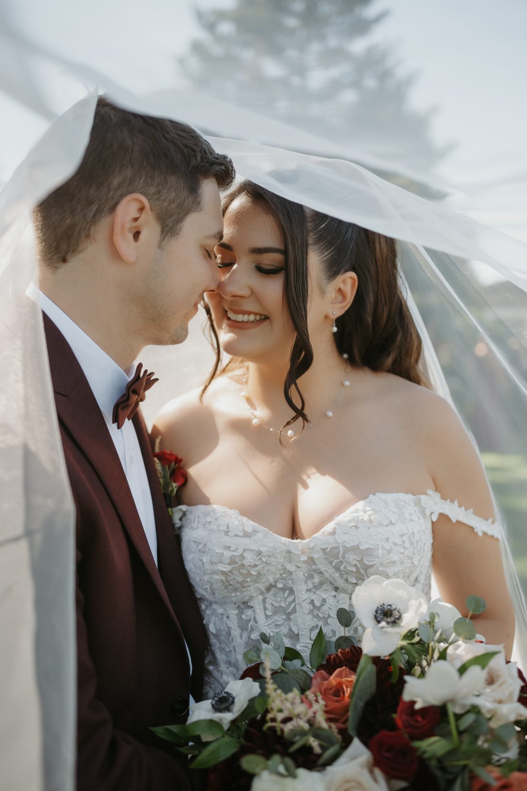Bride and groom standing close under the brides veil, smiling softly while bride holds a bouquet of roses, anemones, dahlias, astilbe and greenery.
