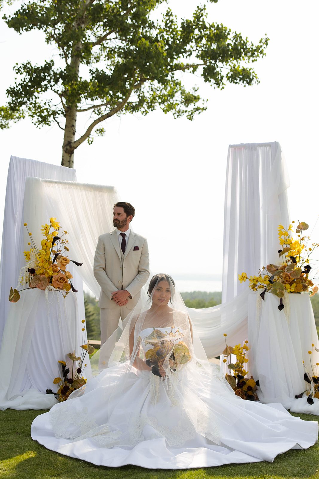 At the ceremony with big panels of white draping fabrics, the bride sits with her veil over her face as she holds her bouquet, her groom behind her, hands clasped looking into the distance.