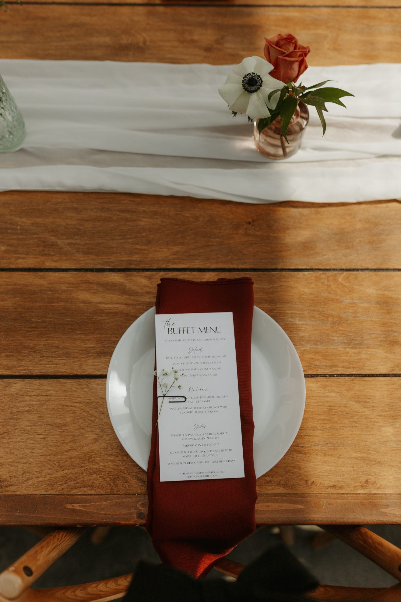 Close up of a wedding place setting featuring a white menu card and burgundy napkin on a white plate, styled on a wooden table with soft white runner.