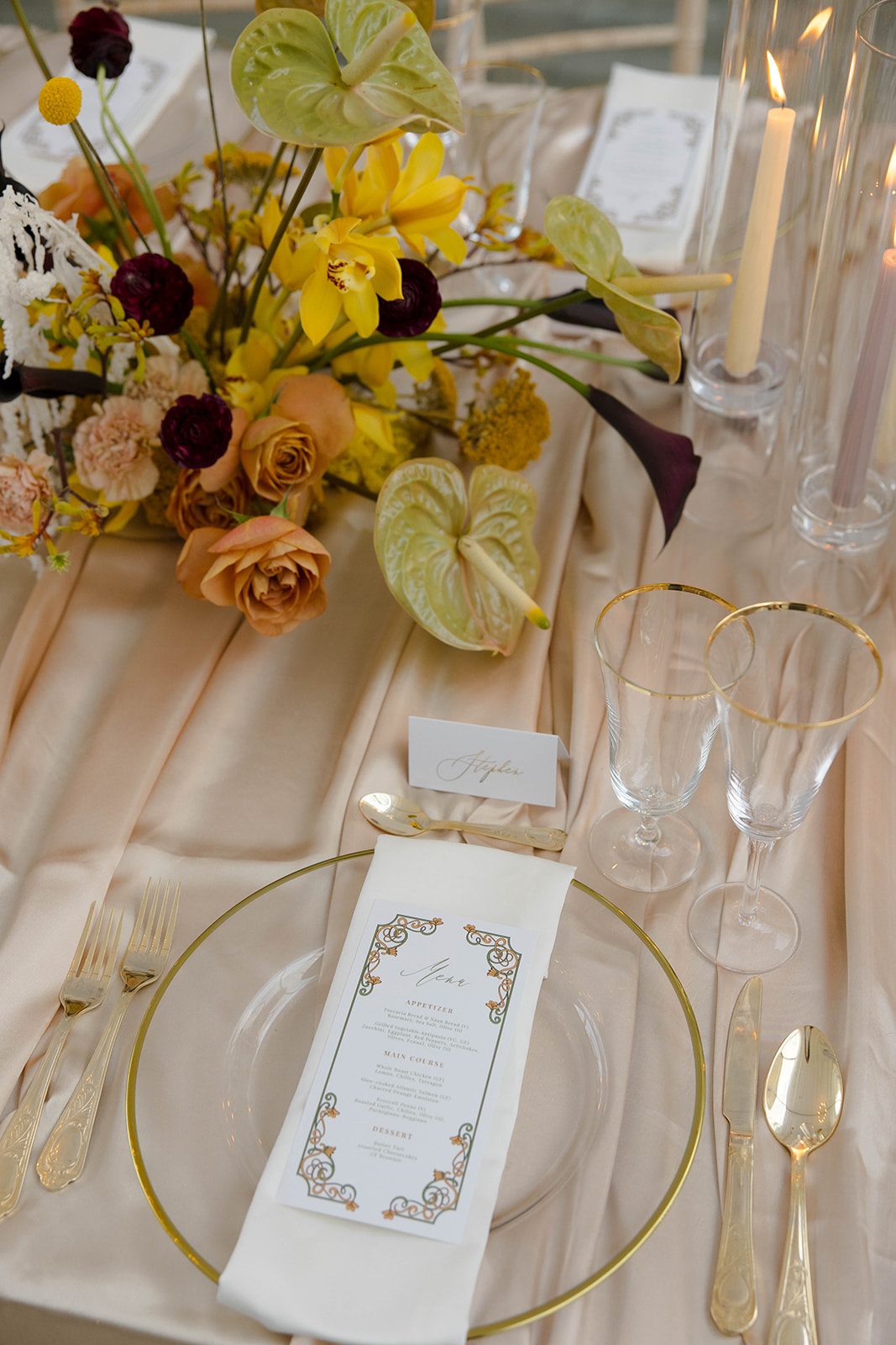 A capture of the place settings with layers of champagne satin on the table, beautiful bold centrepiece, gold rimmed glassware and charger plates with beautiful stationery on top of white linens.