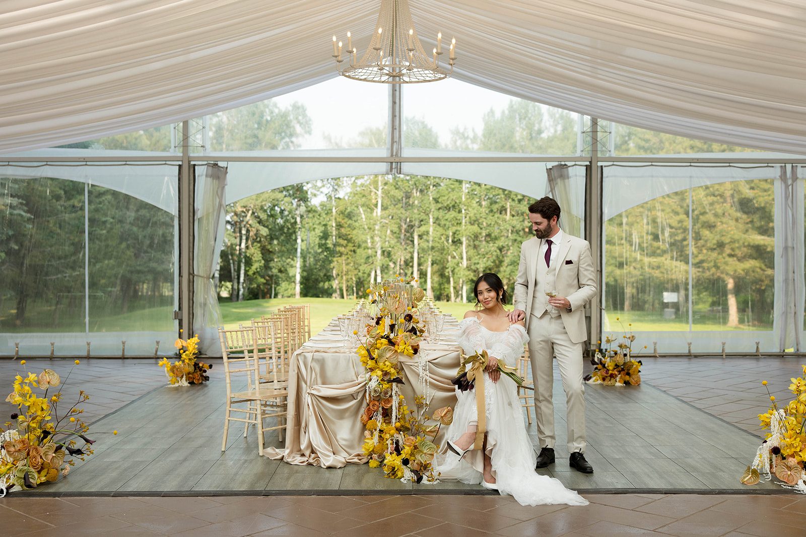 The bride sits next to the luxurious tablescape with her hands and bouquet on her knee while the groom stands beside her, gazing at her lovingly with his hand on her shoulder.