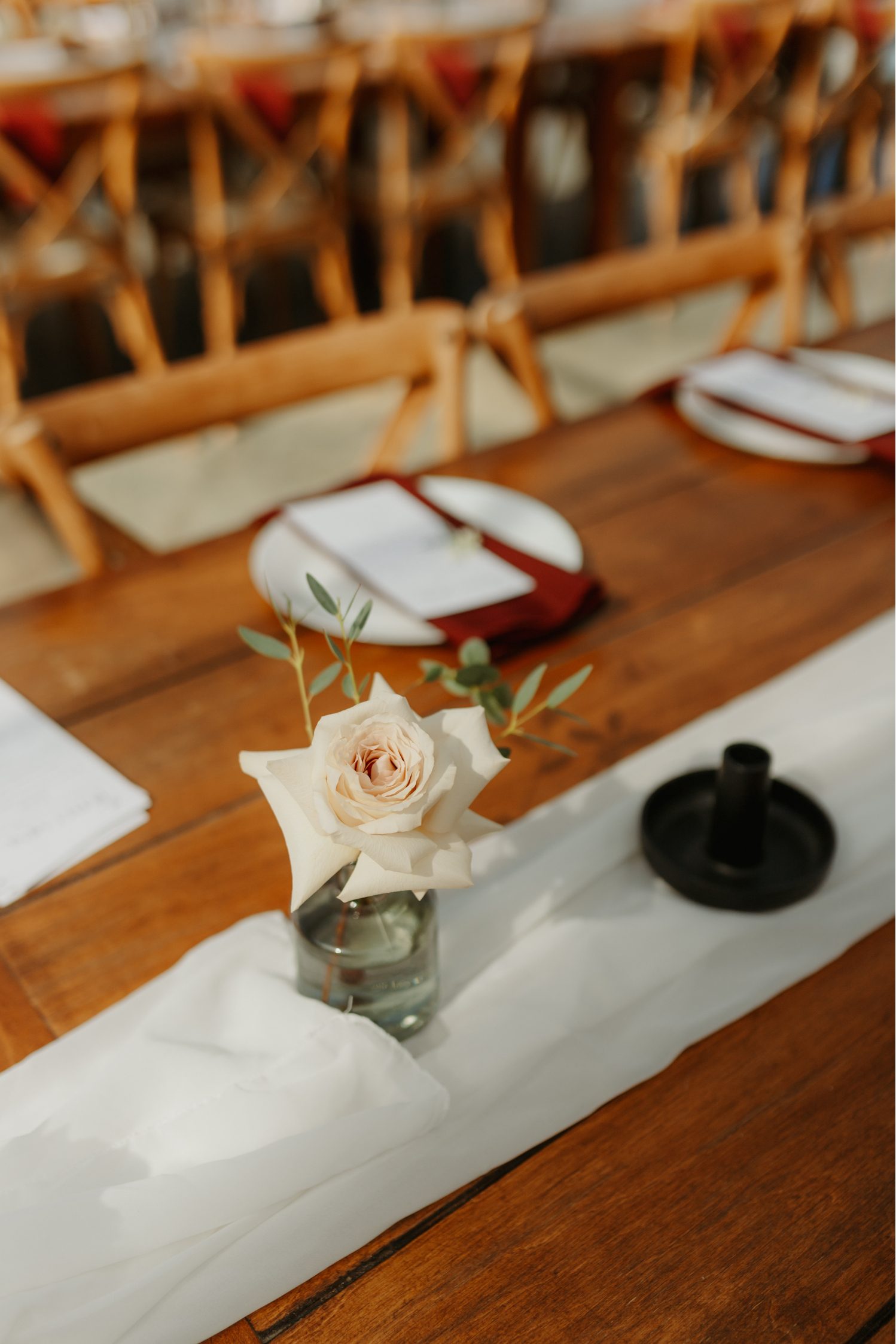 Single blush rose in a small glass bud vase placed on a wooden reception table with a white fabric runner and blurred place settings in the background.