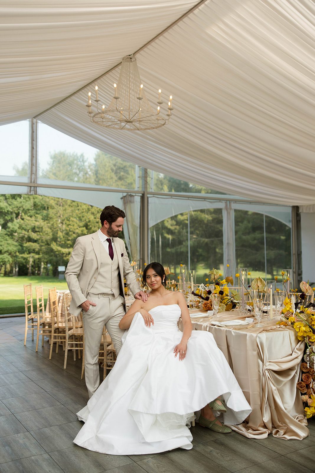 The bride sits in a chair next to the stunning tablescape looking t the camera while the groom stands beside her, gazing at her with his hand resting on her shoulder.