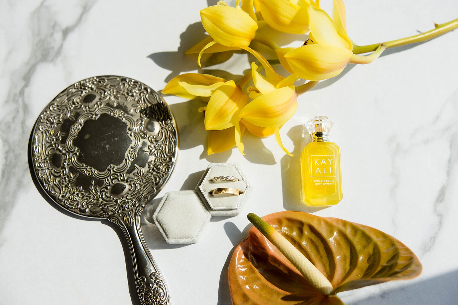 A beautiful, sunny flat lay featuring an antique hand mirror, wedding bands, perfume, bright yellow orchids and a gorgeous tropical anthurium.