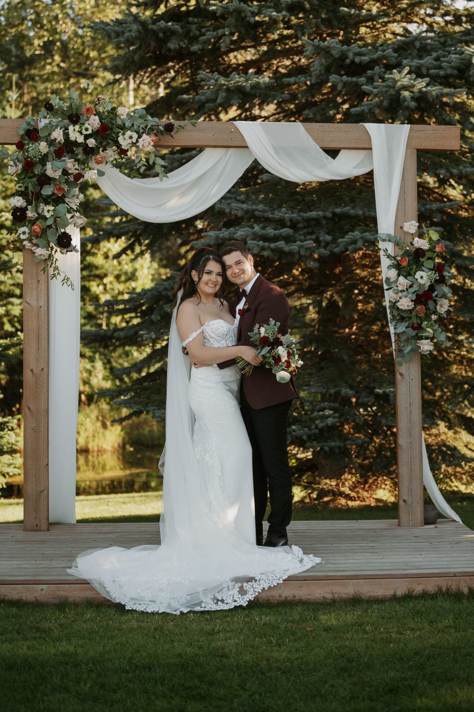 Bride and groom stand embracing beneath their floral adorned wooden ceremony arch surrounded by evergreens.
