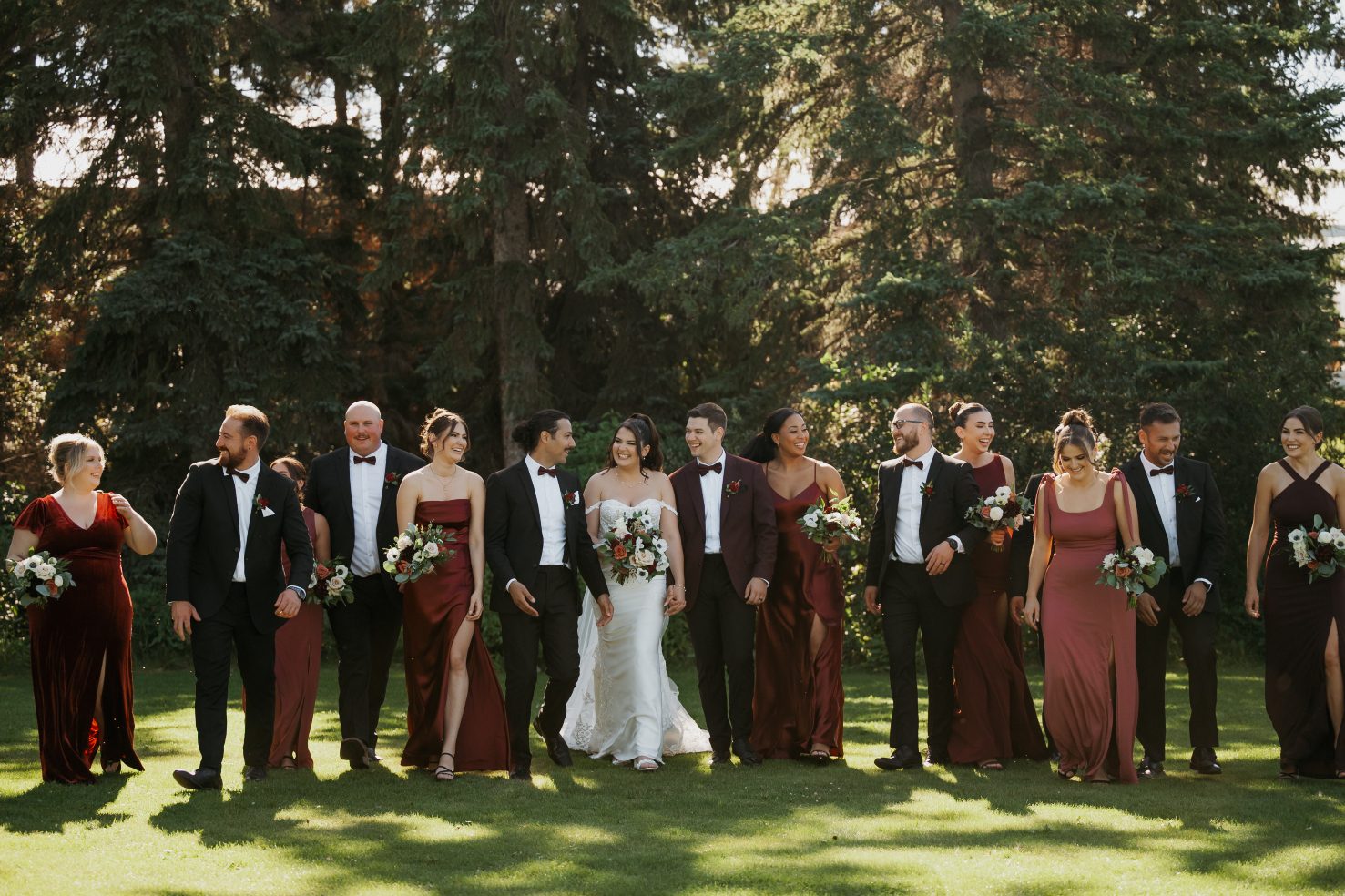Bride and groom walking with their wedding party in mixed burgundy and black attire, sharing relaxed smiles in soft afternoon light
