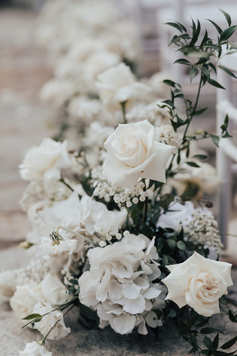 Aisle Arrangements Close up of a white floral aisle arrangement featuring roses, hydrangreas, baby's breath and greenery.
