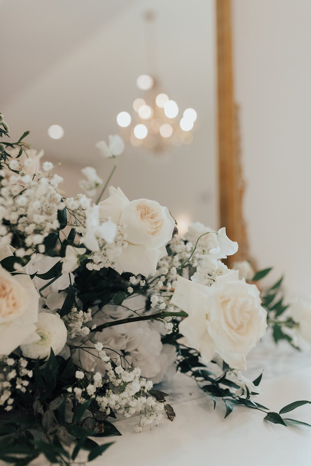 Mr + Mrs Day Close up of white roses, ranunculus and baby's breath arrangement with greenery against a mirror reflecting the image of a chandelier softly blurred.