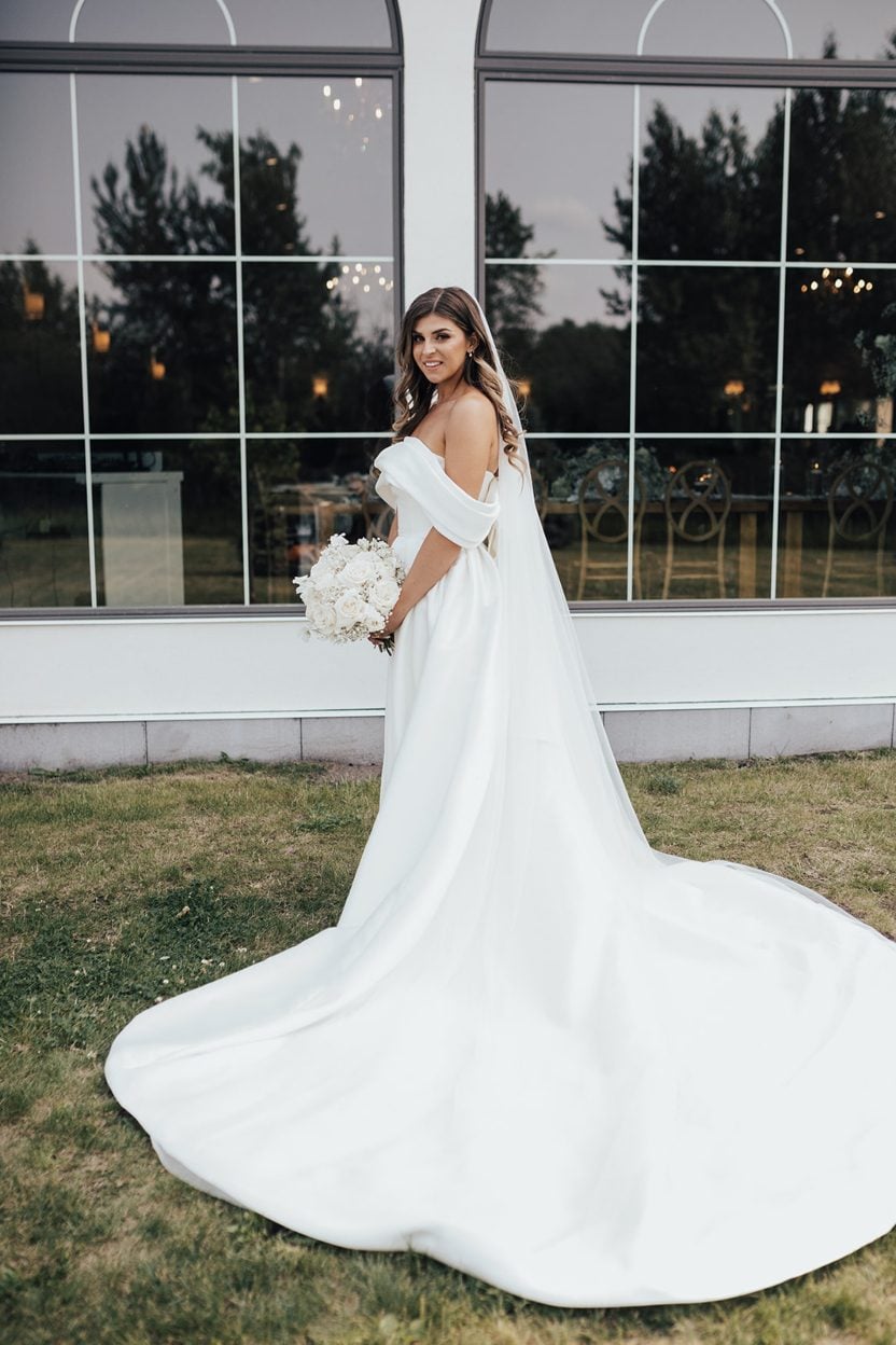 Beautiful Bride Bride standing outdoors in front of arched windows, wearing an of the shoulder satin gown with a long train and holding a white rose bouquet.