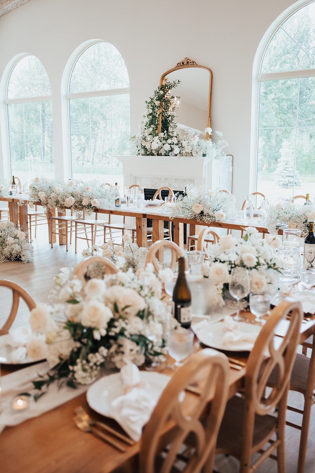 Mr + Mrs Day Wide view of a bright reception space with arched windows, long wooden tables, and lush white floral arrangements.