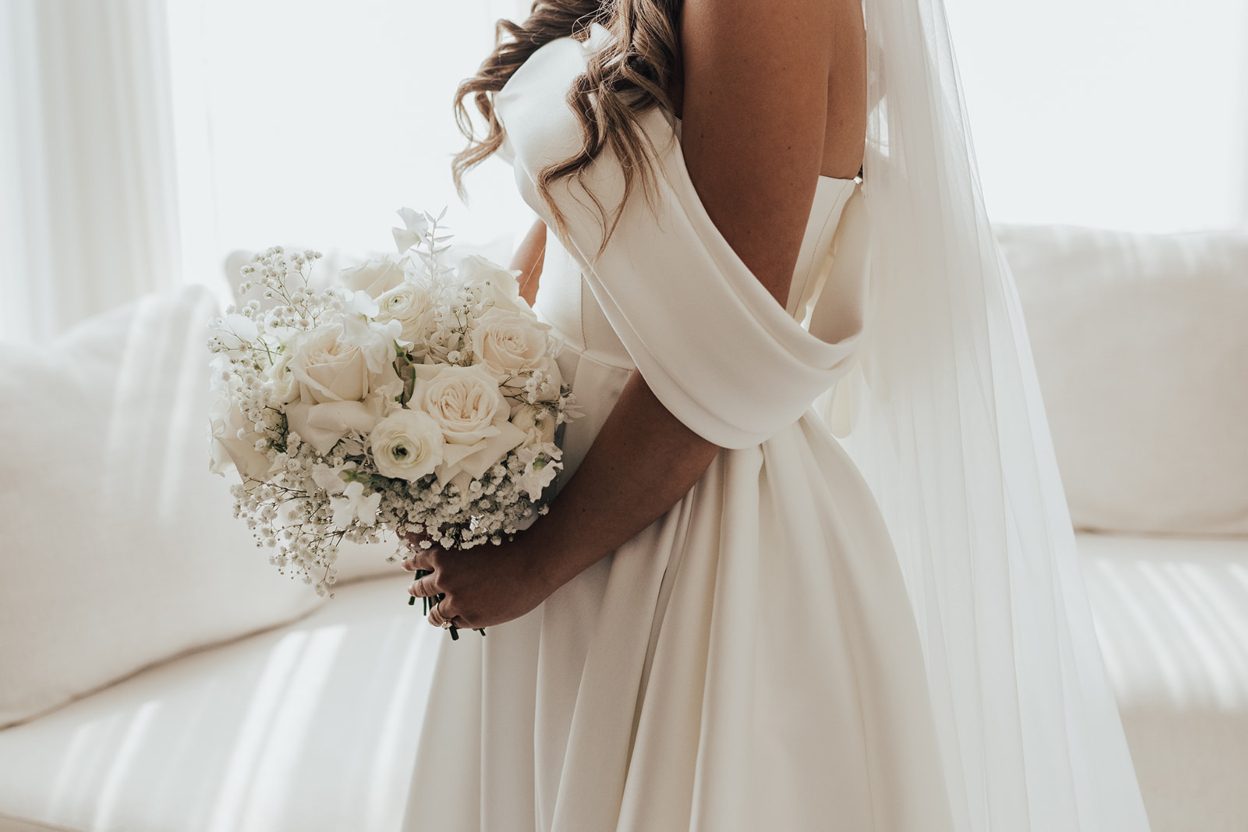 Blushing Bride Close up of the bride holding a white bouquet of roses, ranunculus and baby's breath, showing the draped off the shoulder detail of her satin wedding gown.