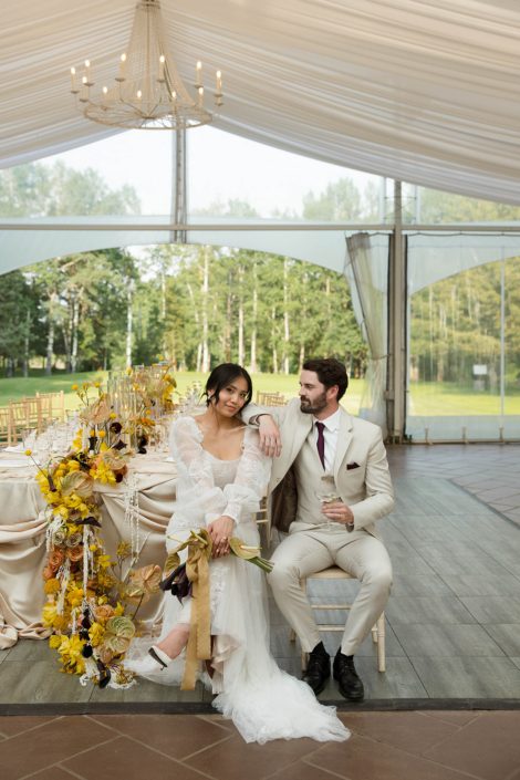 A stunning photoshoot designed with neutrals, mustardy yellow and dark eggplant tones for a bold and beautiful look. Our model couple sits in front of a table with soft satin linens, adorned with yellow and eggplant flowers all the way down, and cascading the end of the table where they sit.