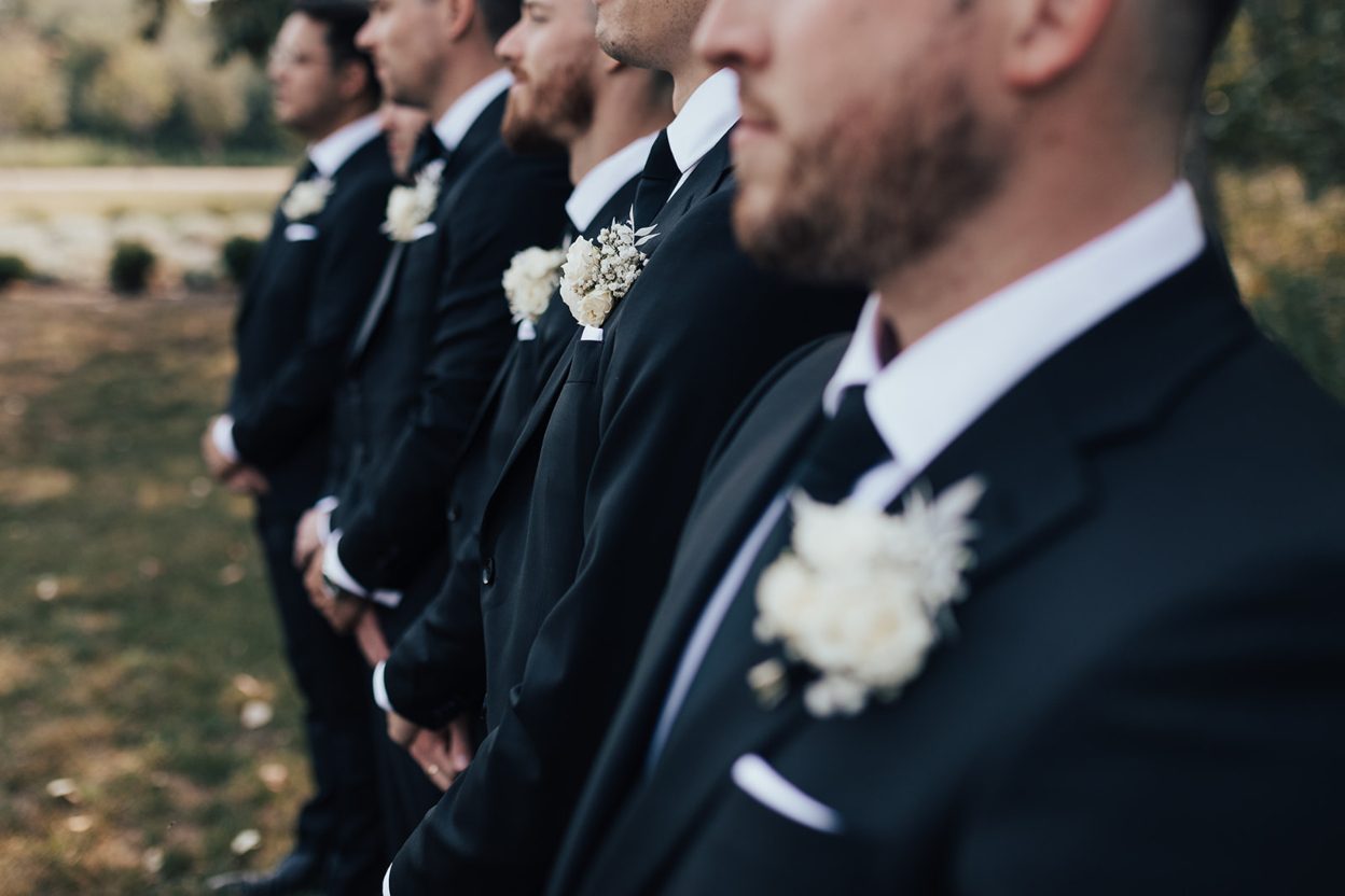Boutonnieres Groomsmen standing in a row wearing black suits and white boutonnieres, photographed in a shallow depth of field lineup.