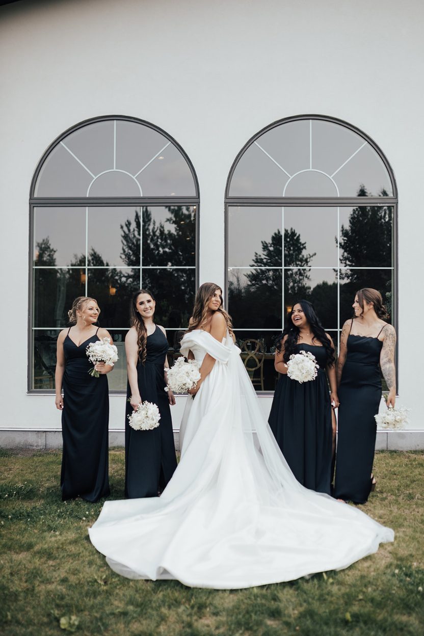 Bride + Bridesmaid Bride and bridesmaids standing outdoors in front of tall arched windows, smiling and holding white bouquets.