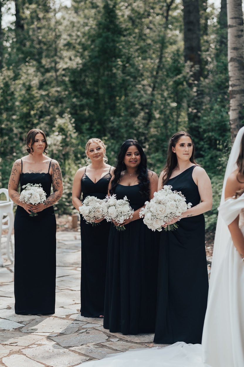 Bridesmaids Bridesmaids standing during the ceremony in black dresses holding white rose and baby's breath bouquets.