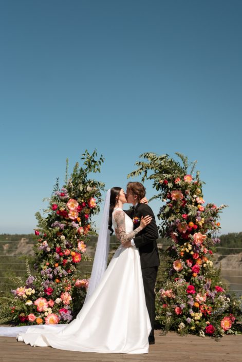A more alternative Calyx bride with tattooed sleeves and her groom share a kiss in front of a broken arch full of pinks, oranges and yellow at an outdoor ceremony with stunning blue skies.