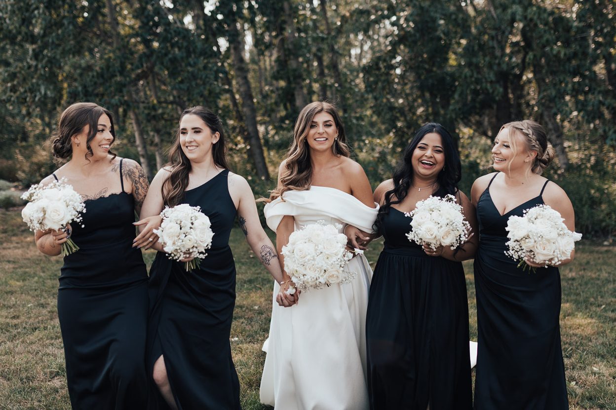 Calyx Bridal Party Bride walking arm in arm with her bridesmaids outdoors, all smiling and holding white bouquets.