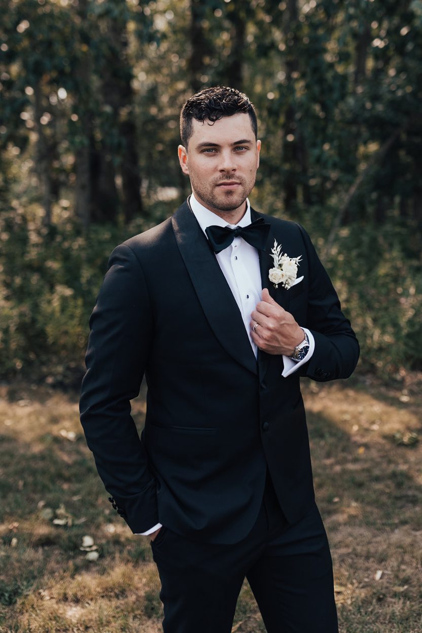 Calyx Groom Groom standing outdoors in a black tuxedo and bow tie, holding his lapel while looking confidently at the camera.