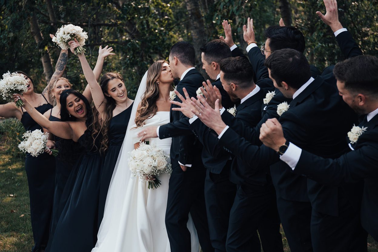 Celebrating Bride and groom kiss while their bridesmaids and groomsmen cheer around them, celebrating outdoors.