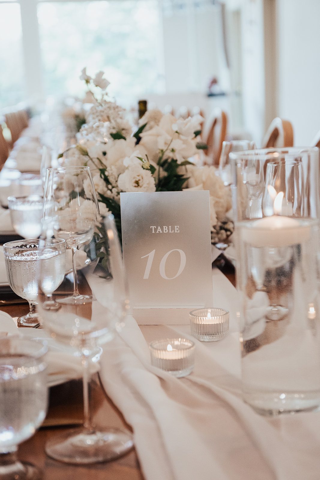 Mr + Mrs Day Table setting featuring a frosted acrylic table number, white floral centrepiece, glassware and glowing votive candles along a soft white fabric runner.