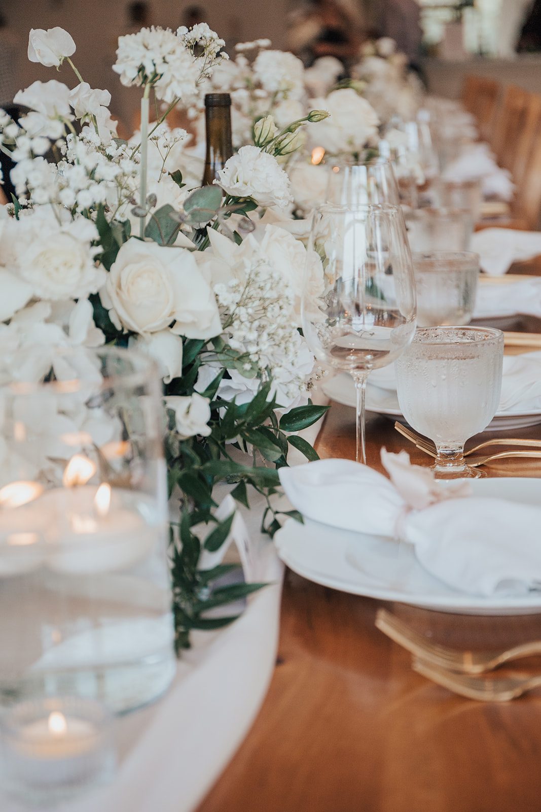 Mr + Mrs Day A row of white centrepieces featuring white roses, hydrangea and greenery displayed down a wooden reception table with glassware and white plates.