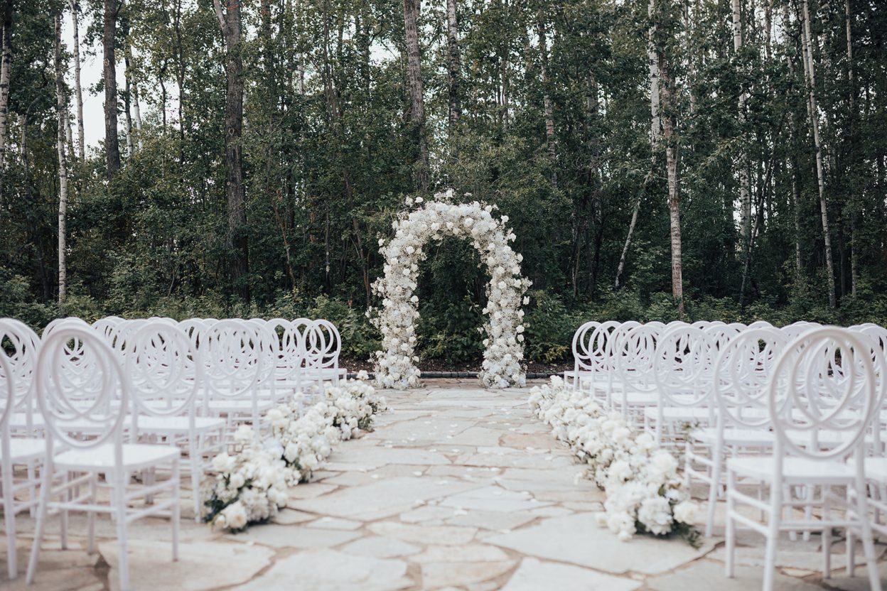 Ceremony Setup Outdoor ceremony set up at Sparrow Lane with white chairs lining the aisle and a lush white floral arch framed by tall forest trees.