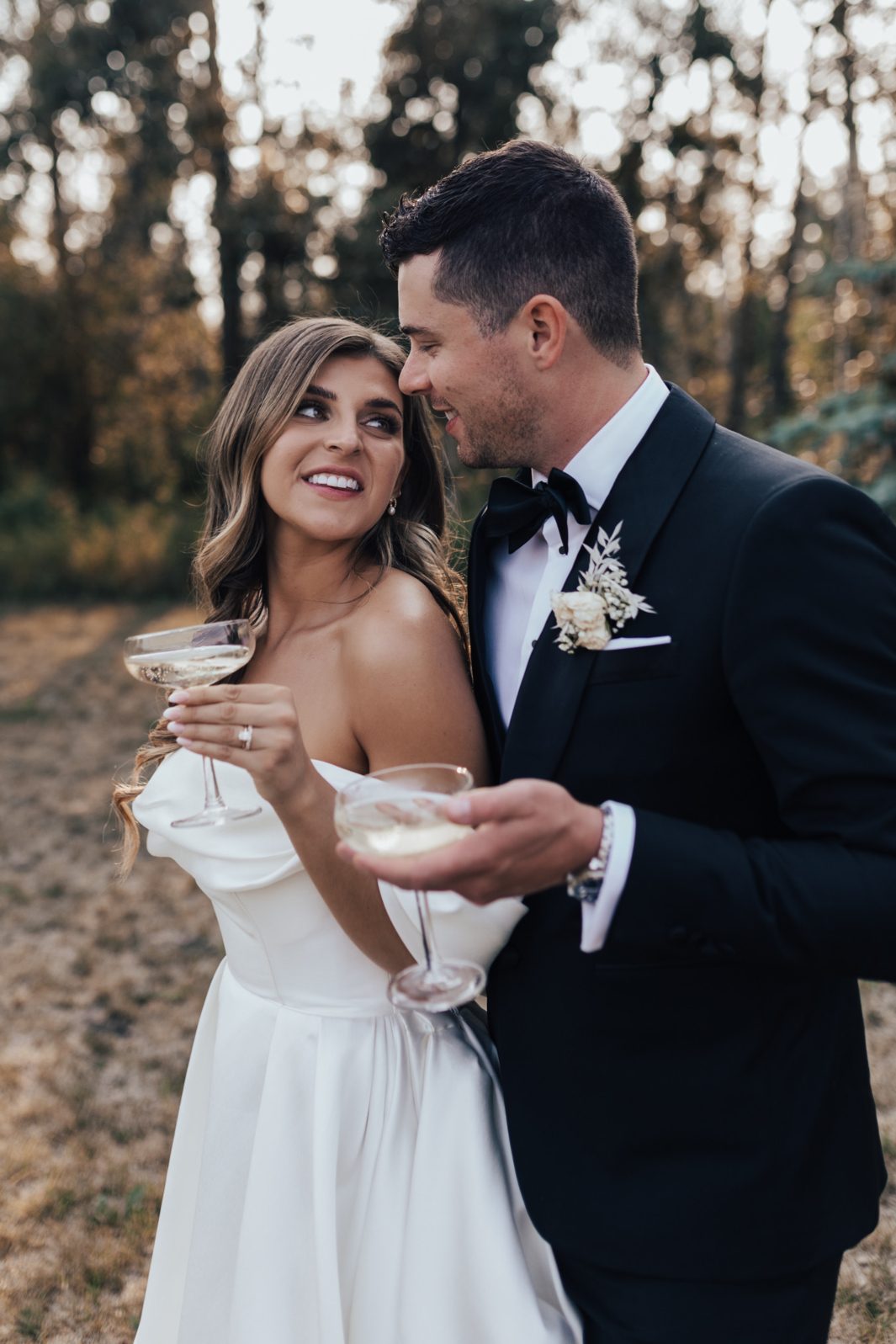 Champagne Bride and groom smiling at each other while holding champagne, standing outdoors in soft evening light.