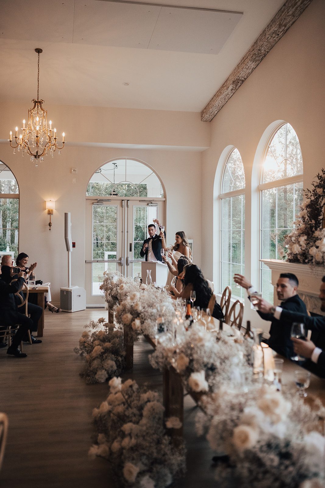 Mr + Mrs Day Guests raising glasses during speeches at the head table, surrounded by abundant white florals and warm natural light.