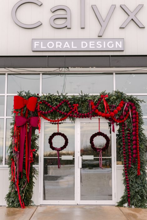 A stunning winter display in front of the store for Christmas with lush noble fir, velvet bobbles in reds and burgundy accented with oversized bows on the left side in matching velvet.
