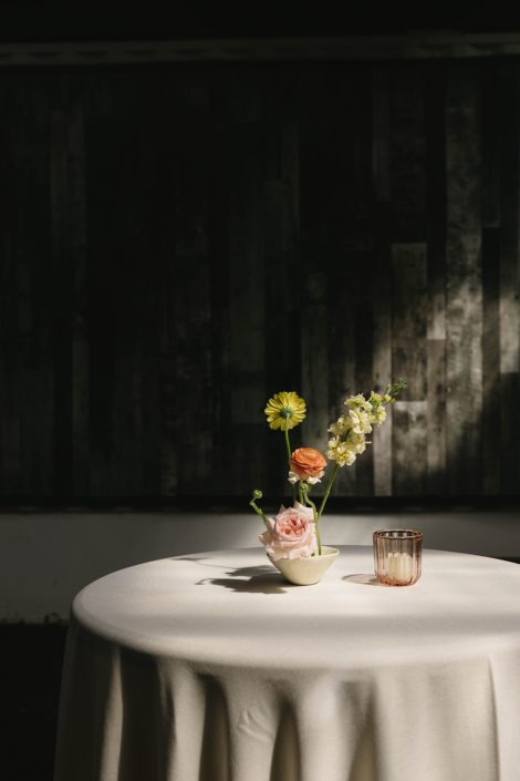 An ikebana inspired centrepiece at a cocktail table featuring ranunculus, stock and a mini Gerbera daisy in a spotlight of natural light from a nearby window.