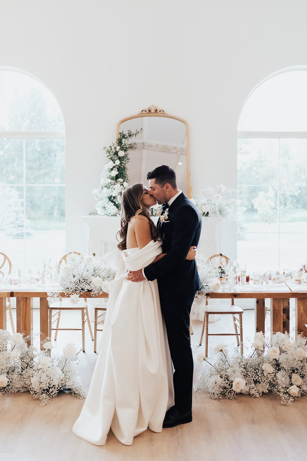 Mr + Mrs Day Bride and groom sharing a kiss in front of their floral filled head table, framed by arched windows and a floral accented mirror.
