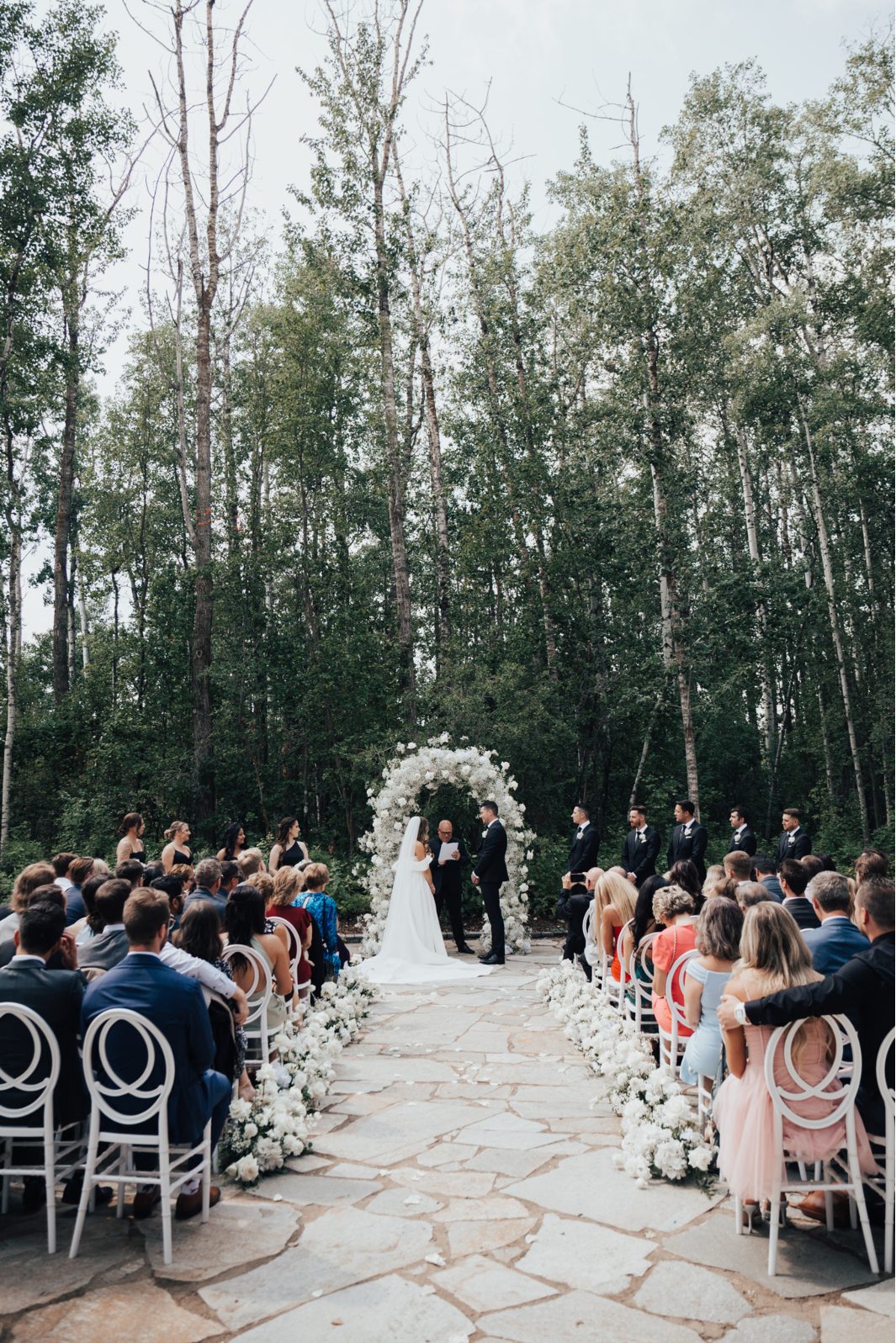 Day Ceremony View of the outdoor ceremony as guests look on and the couple stands beneath a white rose and baby's breath arch.