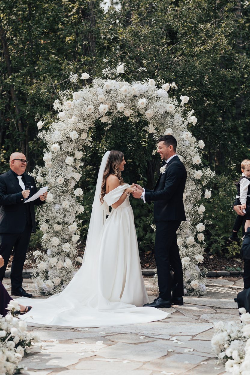 During Ceremony Bride and groom holding hands while exchanging vows beneath a full white floral arch.