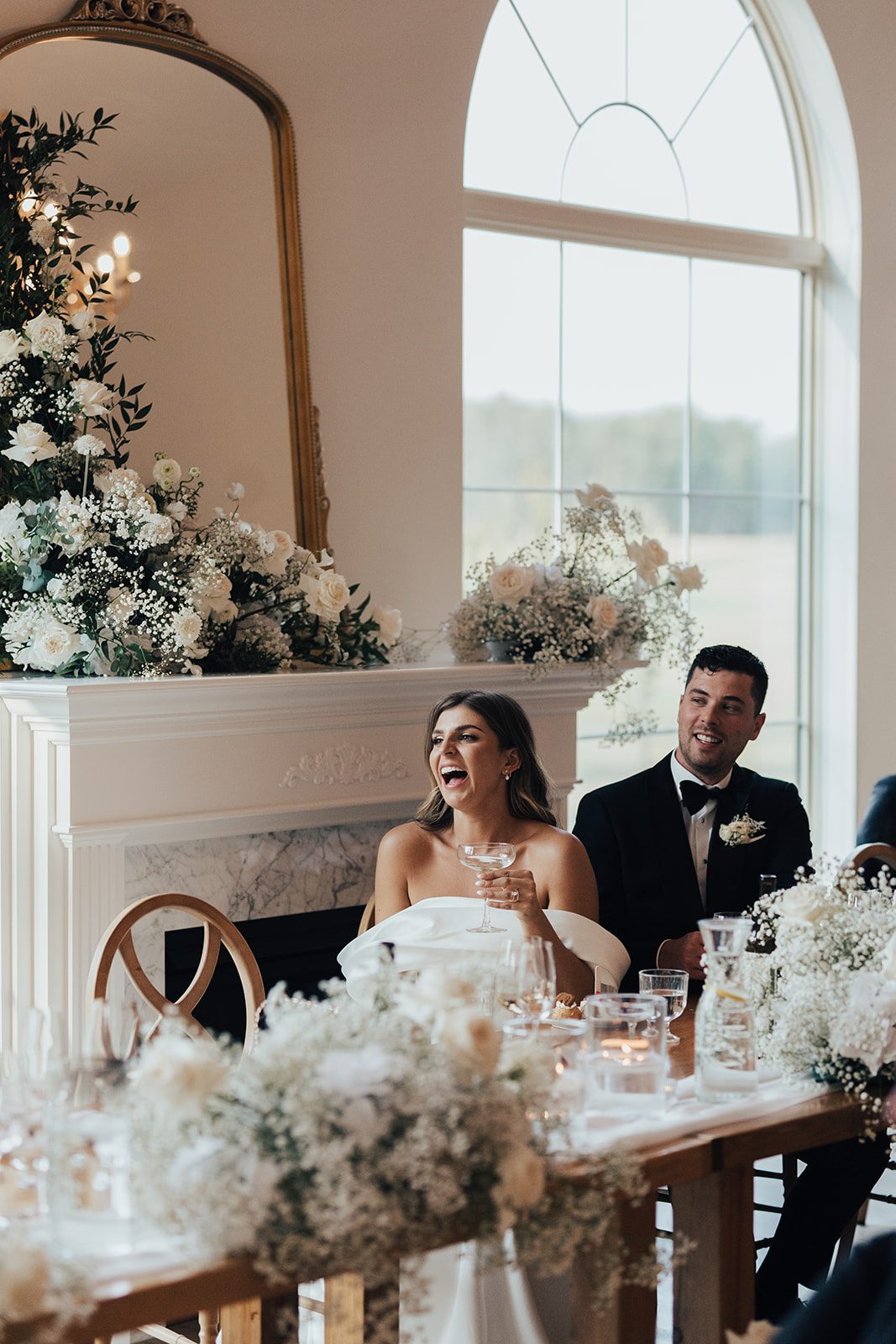 Mr + Mrs Day Bride laughing while holding a champagne as the groom smiles beside her, seated at the floral decorated head table.