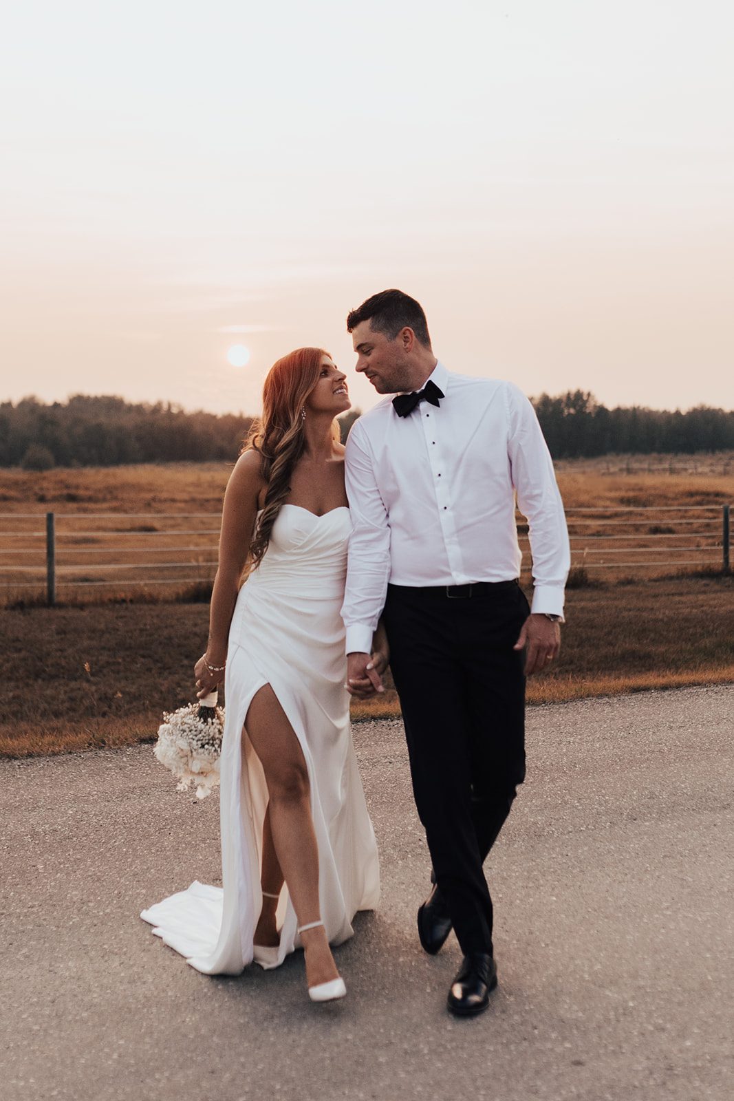 Mr + Mrs Day Bride and groom walking hand in hand at sunset, with the bride holding a white bouquet and the sky glowing softly behind them.