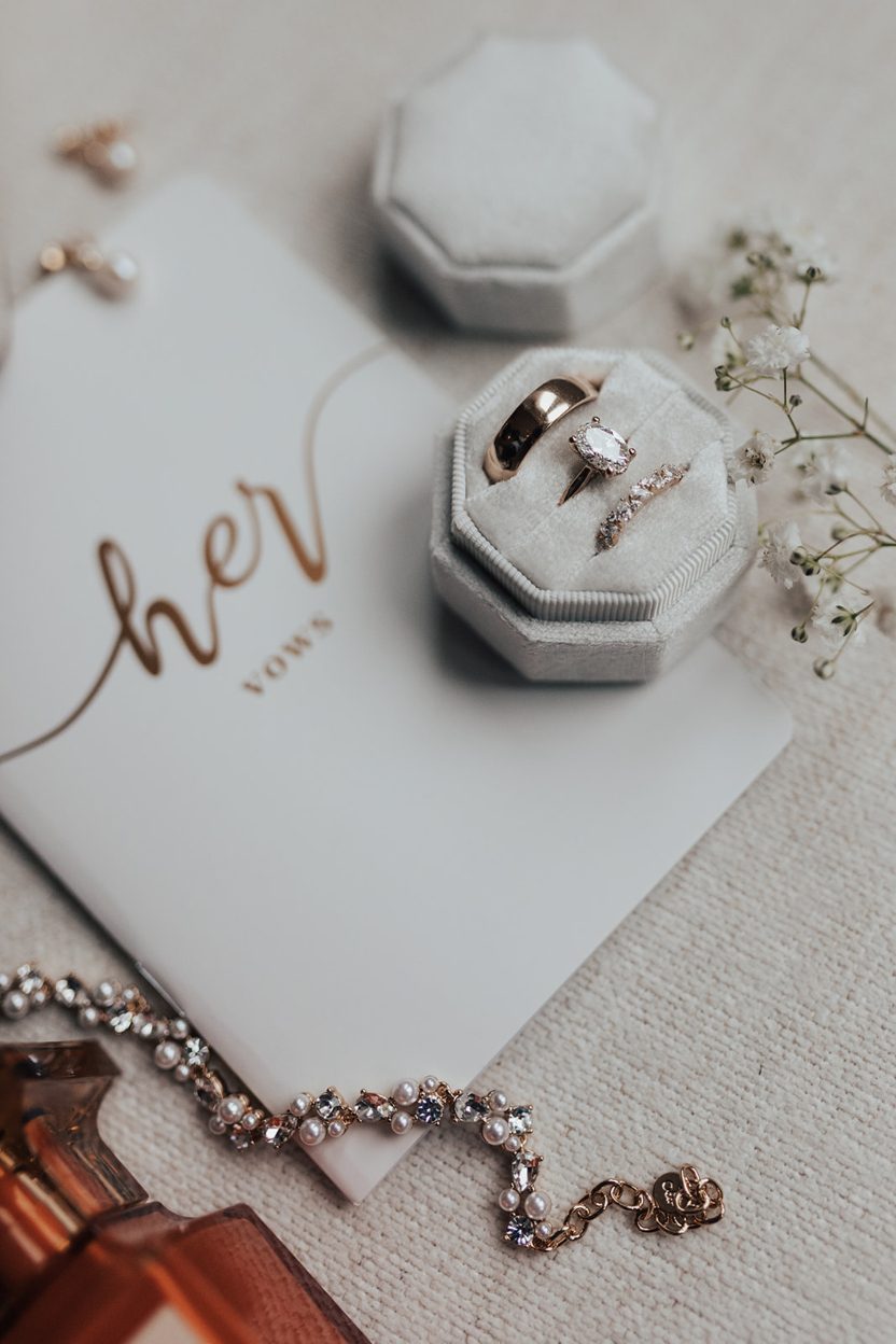 Flat Lay Flatlay of wedding details including a vow booklet, ring box with the bridal set, a gold band, pearl bracelet and a perfume bottle.