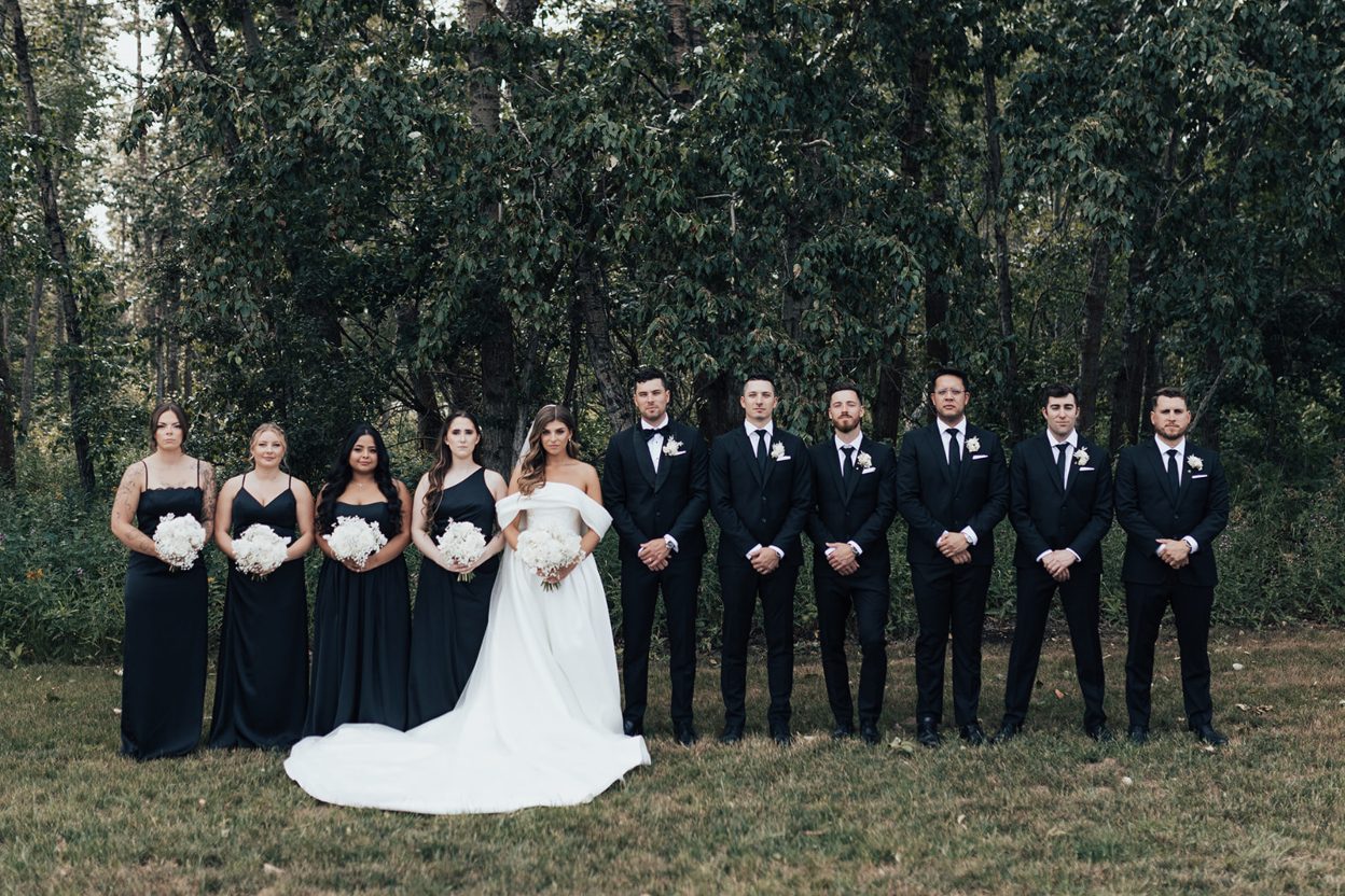 Formal Wedding Party Wedding party lined up outdoors with the bride and groom centred. Bridesmaids to the left wearing black dresses with white bouquets and the groomsmen to the right in black suits with white boutonnieres.