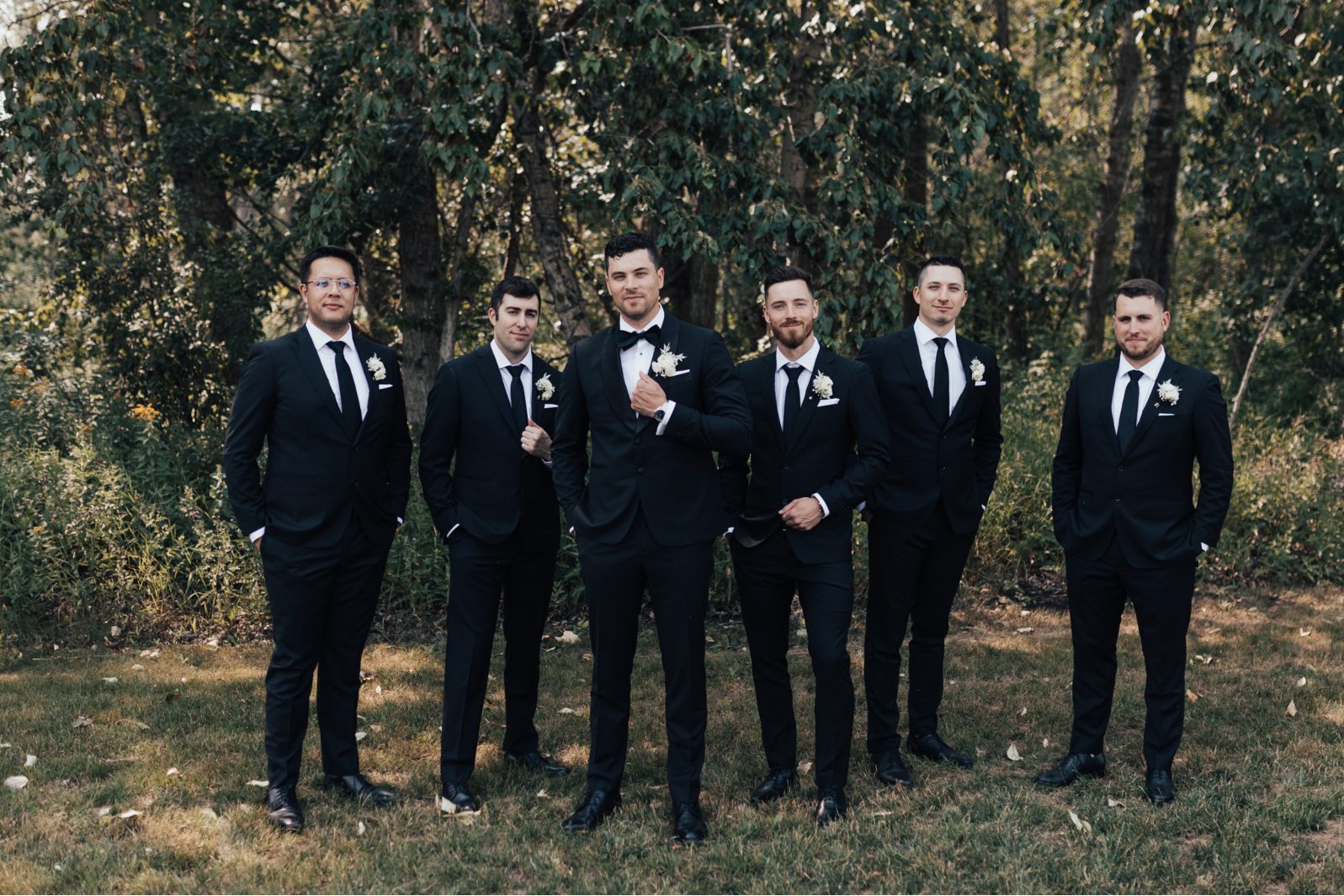 Groom + Groomsmen Groom standing with his groomsmen outdoors, all wearing black suits and white boutonnieres.