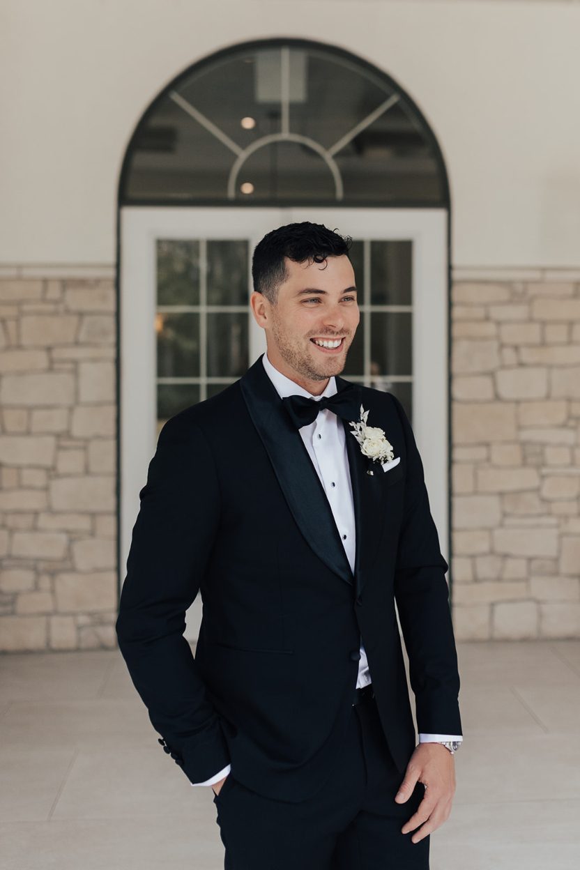 Groom Groom smiling while standing in front of an arched window, dressed in a black tuxedo and sporting a white boutonniere.