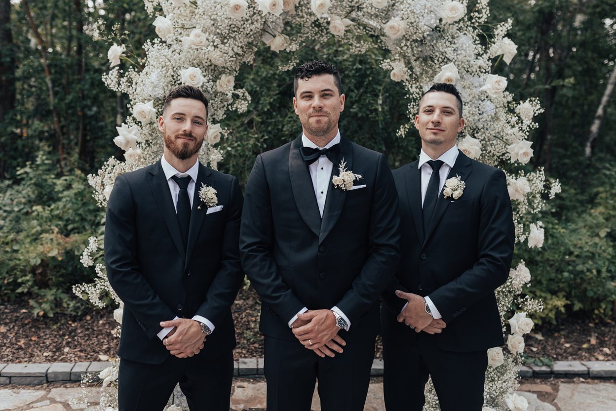 Groomsmen Groom standing with two groomsmen in front of a white rose and baby's breath arch, all wearing black suits with matching white boutonnieres.