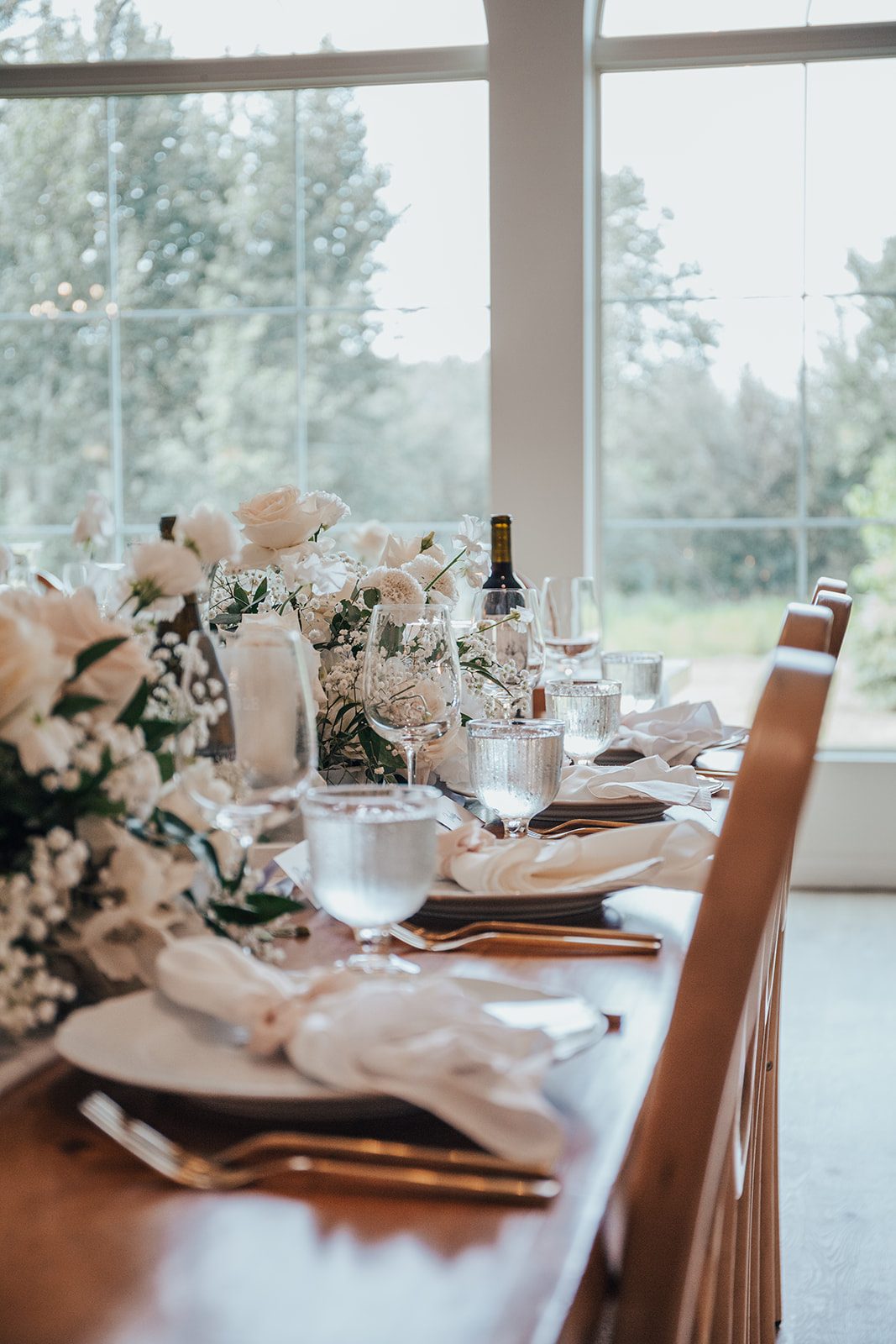 Mr + Mrs Day Close up of an elegant wedding reception table featuring roses, baby's breath and lush greenery arranged along a wooden table with gold flatware, crystal water goblets and soft natural light streaming through large windows.